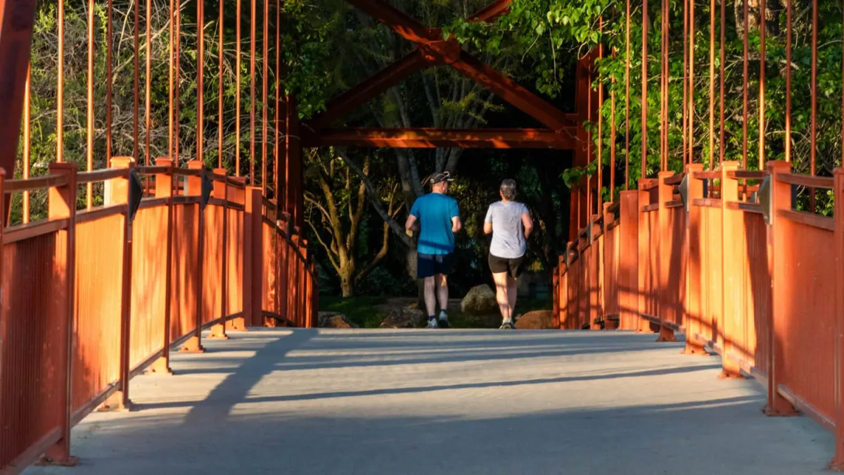 Two people jogging across the Red Bridge on the Boise River Greenbelt, framed by its red railings and green trees