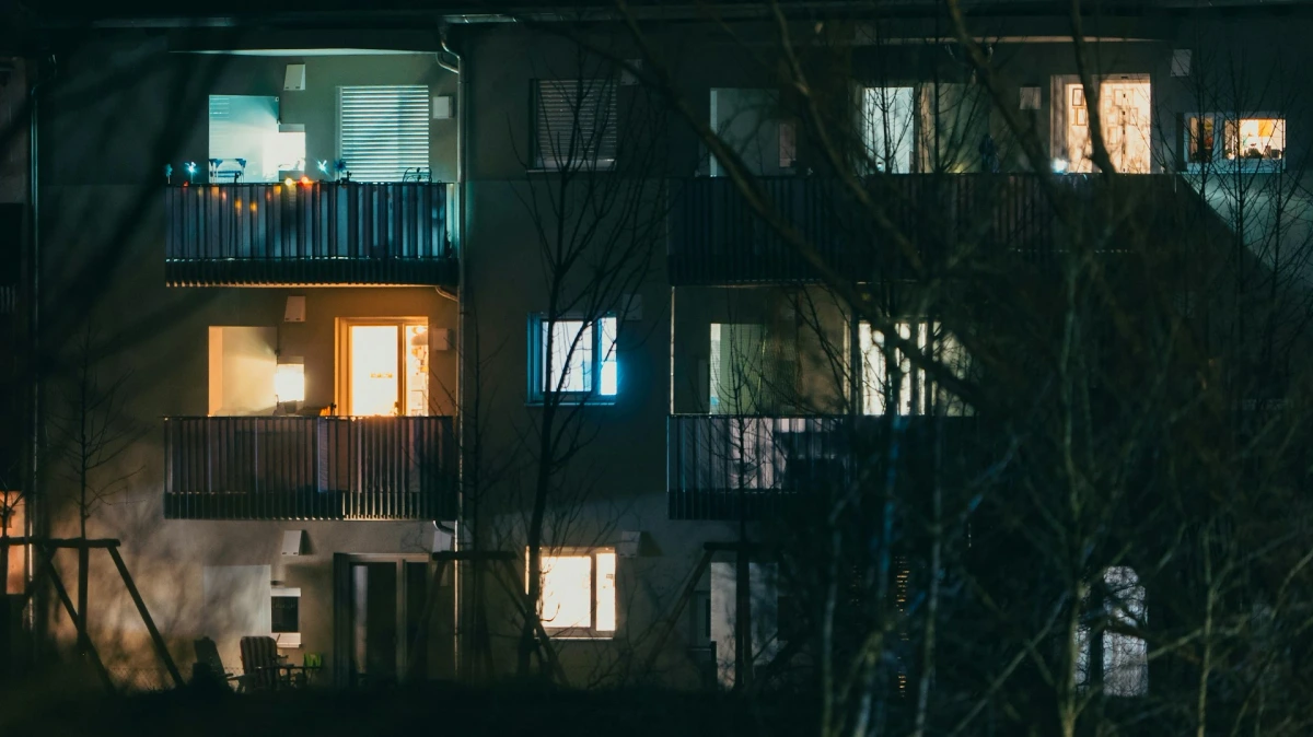 Multi-story apartment building at night, with illuminated windows and balconies partially obscured by bare trees.