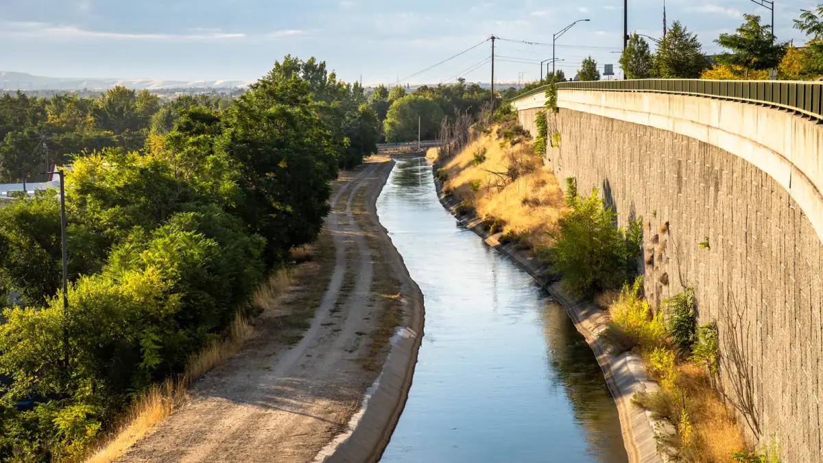 An irrigation canal running through Boise beside mature trees, a maintenance road, and a retaining wall.
