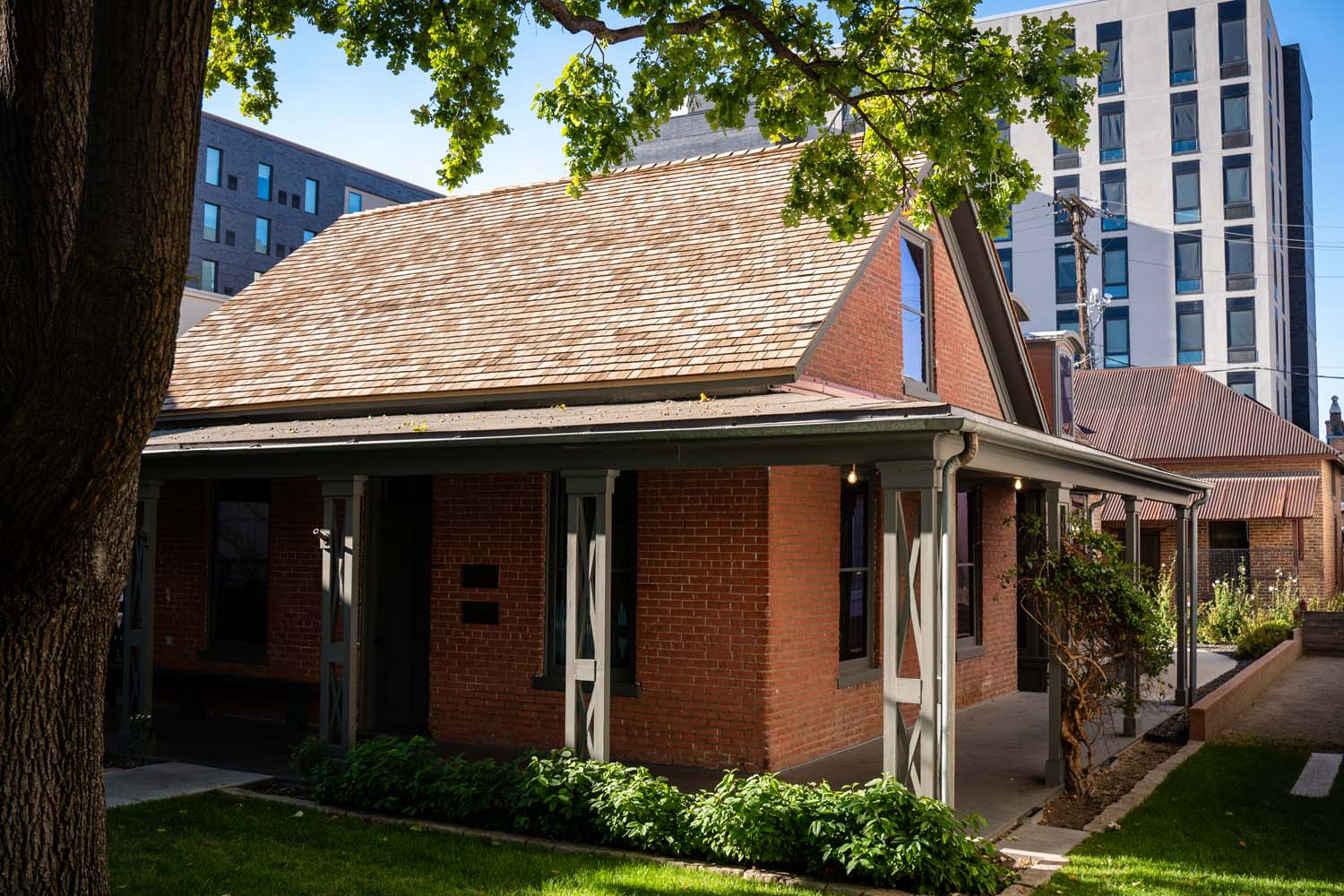 A historic brick house in downtown Boise stands beneath mature trees, surrounded by newer buildings.