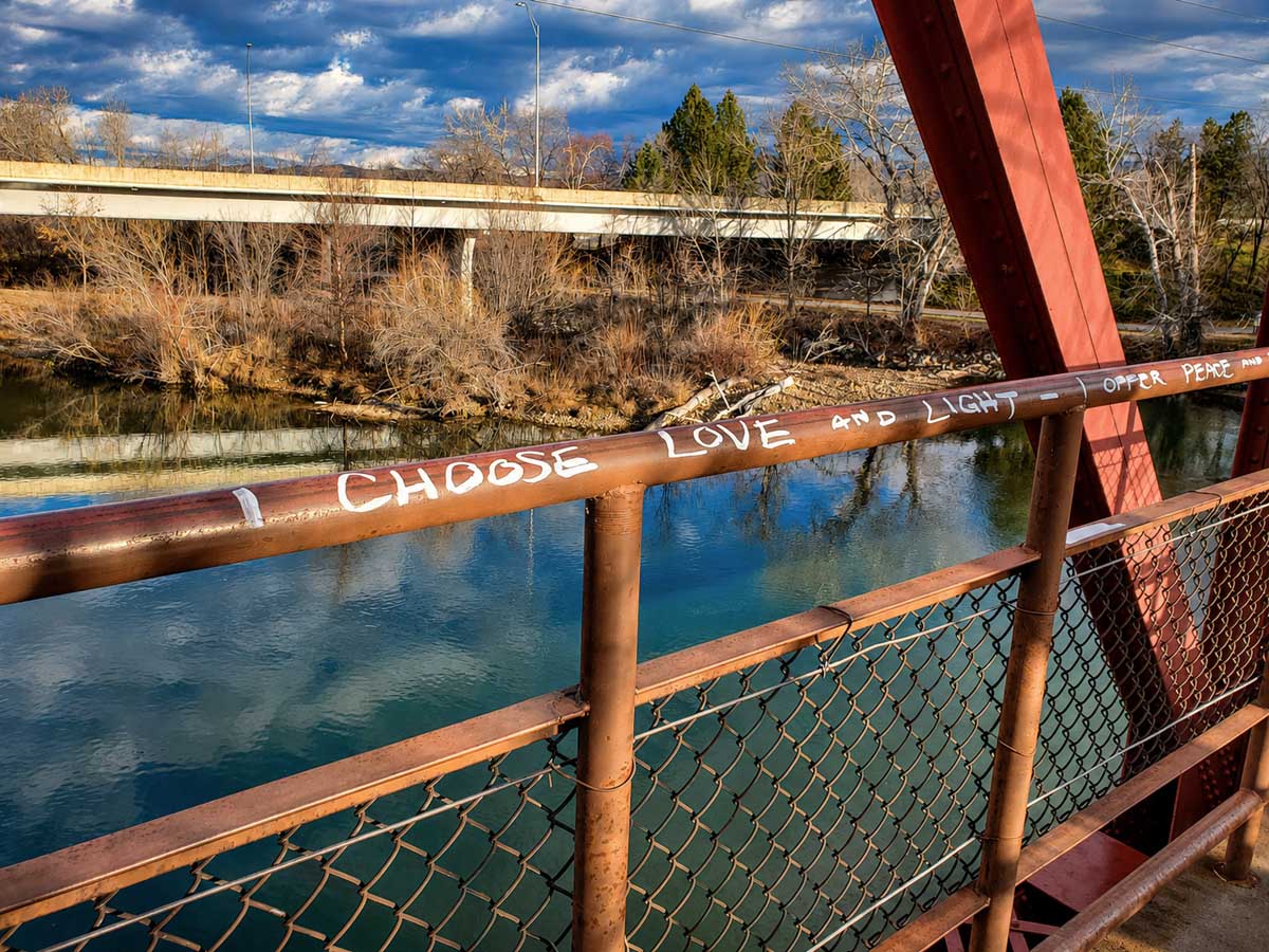 White graffiti on a metal bridge railing reads “I choose love and light,” with the Boise River and trees in the background.