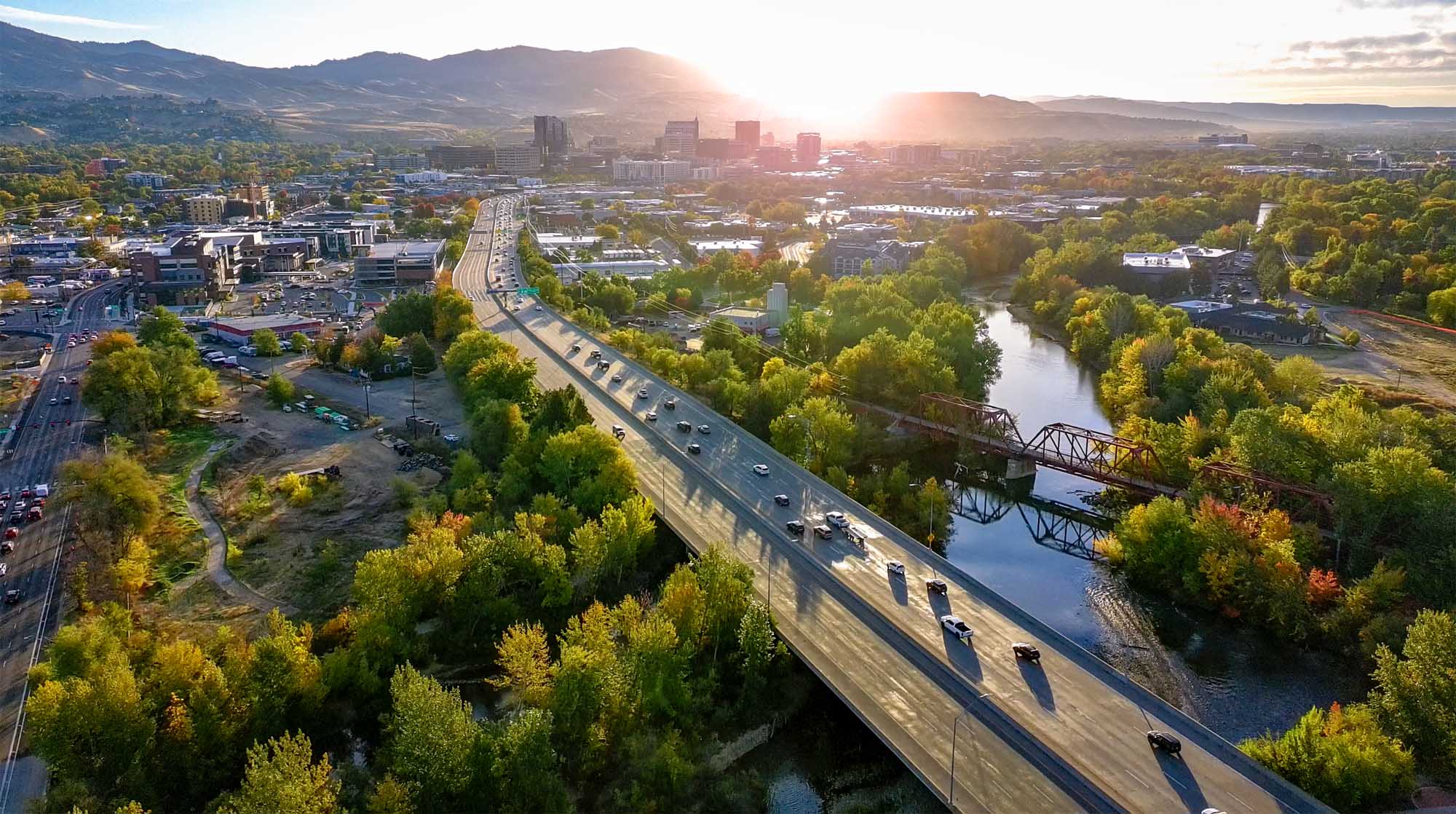 Aerial view of Boise, Idaho at sunrise showing cars on the connector overpass over the river with the downtown skyline and foothills in the distance.
