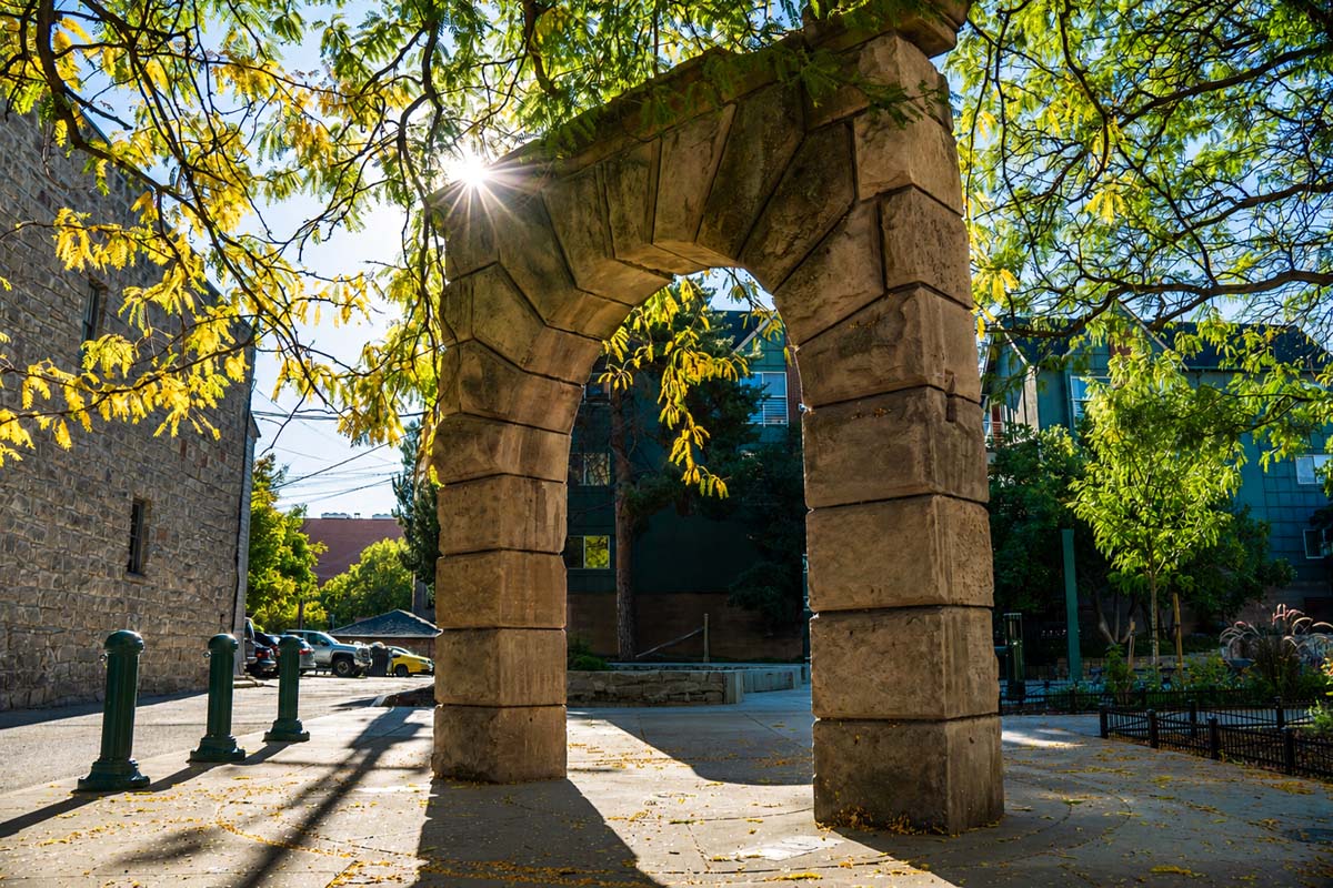 Sunlit photograph of a weathered stone archway in downtown Boise, framed by leafy tree branches and long shadows, with a bright starburst of light filtering through the canopy and nearby buildings and sidewalk visible in the background.