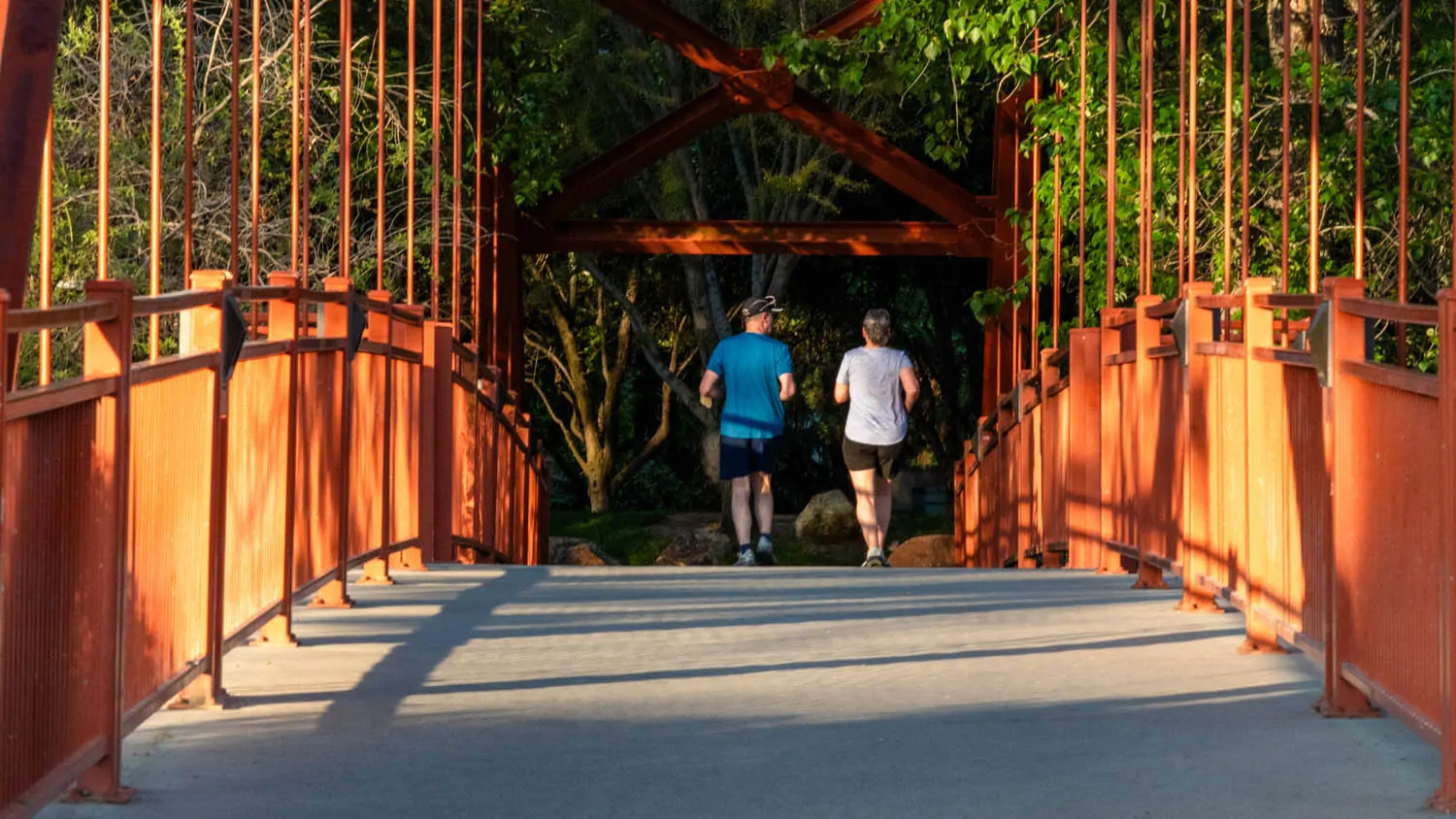 Two people jogging across the Red Bridge on the Boise River Greenbelt, framed by its red railings and green trees