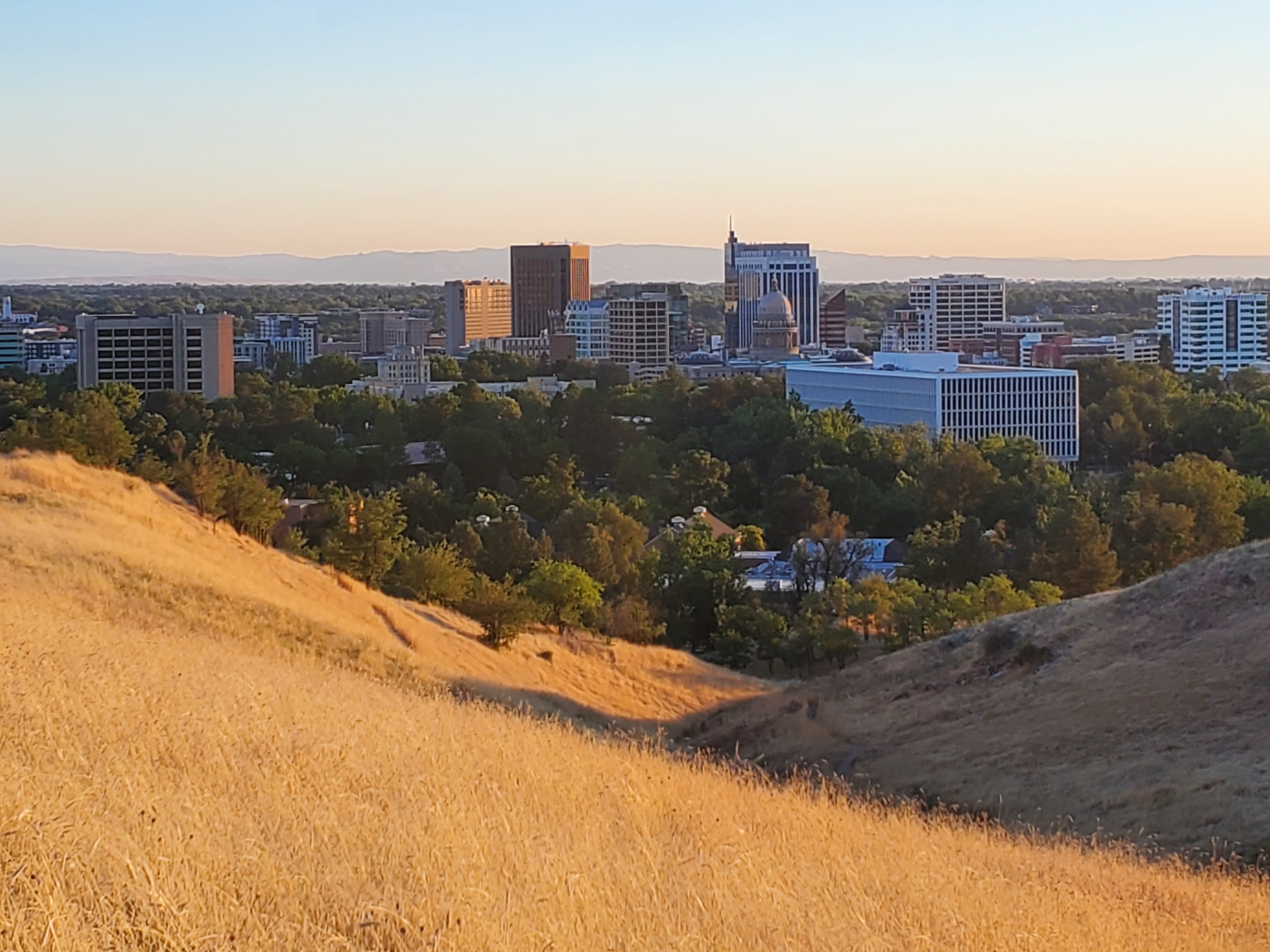 Downtown Boise beyond golden foothills under a pale evening sky