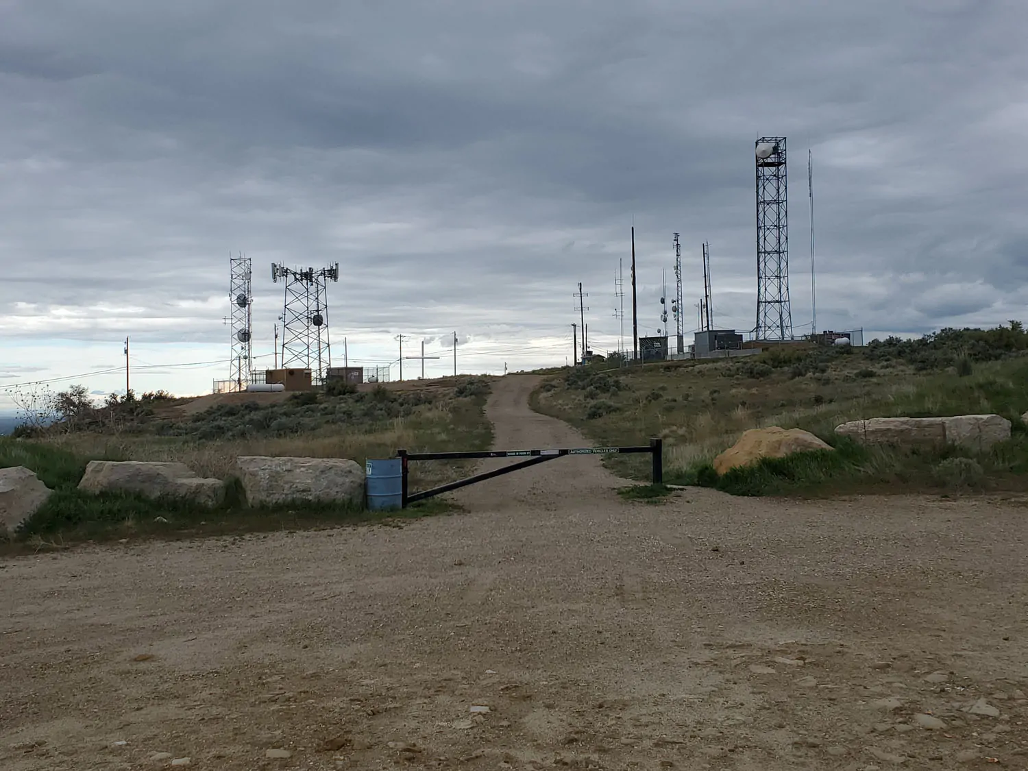 Current Table Rock summit approach with a blocked gravel road, utility towers, power lines, fencing, and other service infrastructure dominating the landscape beneath an overcast sky.