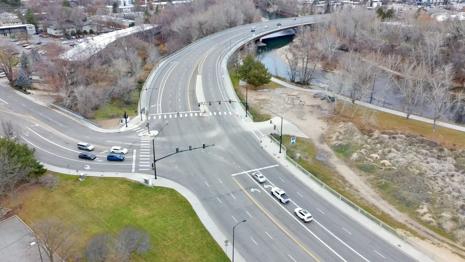 An aerial photograph of the Parkcenter bridge in Boise, Idaho, showing a multi-lane asphalt roadway and heavy concrete infrastructure crossing the Boise River, surrounded by utilitarian traffic lights and winter-toned vegetation.